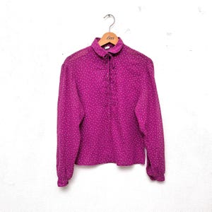 Peut inclure: Une blouse rose à manches longues avec un motif de pois blancs. La blouse a un nœud papillon au cou et un devant boutonné.