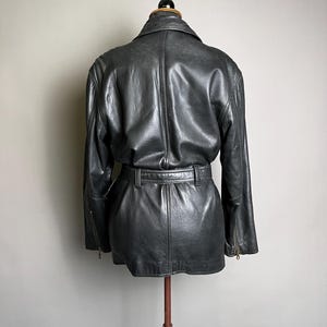 May include: A dark gray leather jacket with a belted waist and long sleeves. The jacket has a classic design with a collar and zipper details on the sleeves. The jacket is displayed on a mannequin.