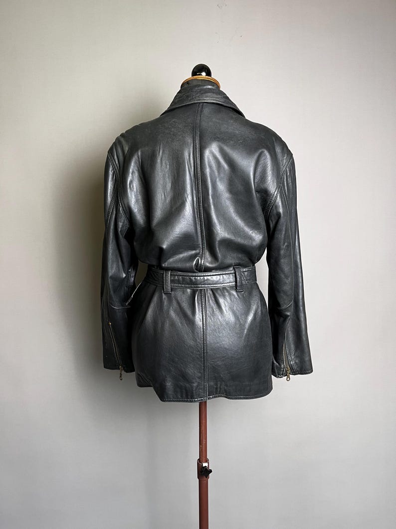 May include: A dark gray leather jacket with a belted waist and long sleeves. The jacket has a classic design with a collar and zipper details on the sleeves. The jacket is displayed on a mannequin.
