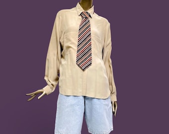 Könnte beinhalten: Ein beiges Langarmhemd mit spitzem Kragen und zwei Brusttaschen, kombiniert mit einer Krawatte mit diagonalen Streifen in Schwarz, Weiß und Rot. Das Outfit umfasst eine hellblaue Jeansshorts, die an einem Schaufensterpuppen-Torso getragen wird.