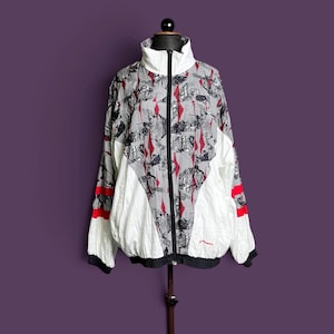 May include: A white, black, and red zip-up windbreaker jacket featuring a high collar, long sleeves with red stripes, and a graphic pattern of black and white shapes with red diamond accents across the chest and back panels.