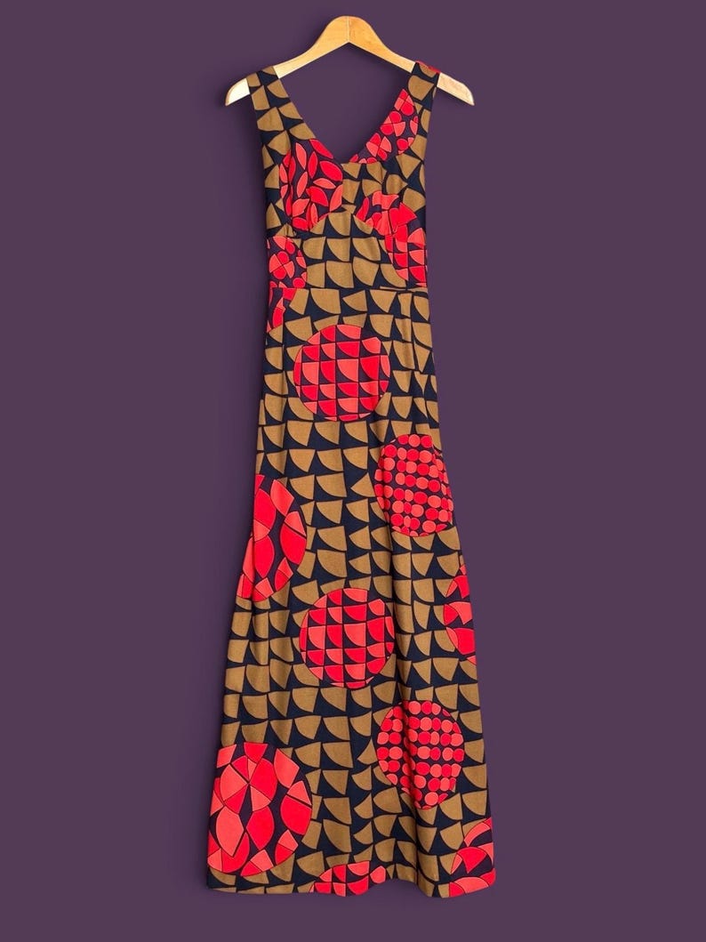 May include: A sleeveless floor-length dress featuring a V-neckline and a repeating geometric pattern. The design consists of dark navy blue semicircles on a tan background, accented by large circular motifs filled with bright red and pink segments.