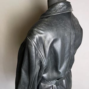 May include: A black leather jacket with a classic design, displayed on a mannequin. The jacket features a collar, visible stitching, and a belt detail at the waist. The leather has a slightly worn appearance.