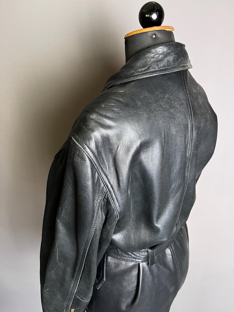 May include: A black leather jacket with a classic design, displayed on a mannequin. The jacket features a collar, visible stitching, and a belt detail at the waist. The leather has a slightly worn appearance.