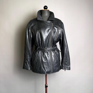 May include: A dark gray leather jacket with a belted waist and an asymmetrical zipper closure. The jacket features a wide collar, multiple zippered pockets, and a classic biker style. The jacket is displayed on a mannequin.