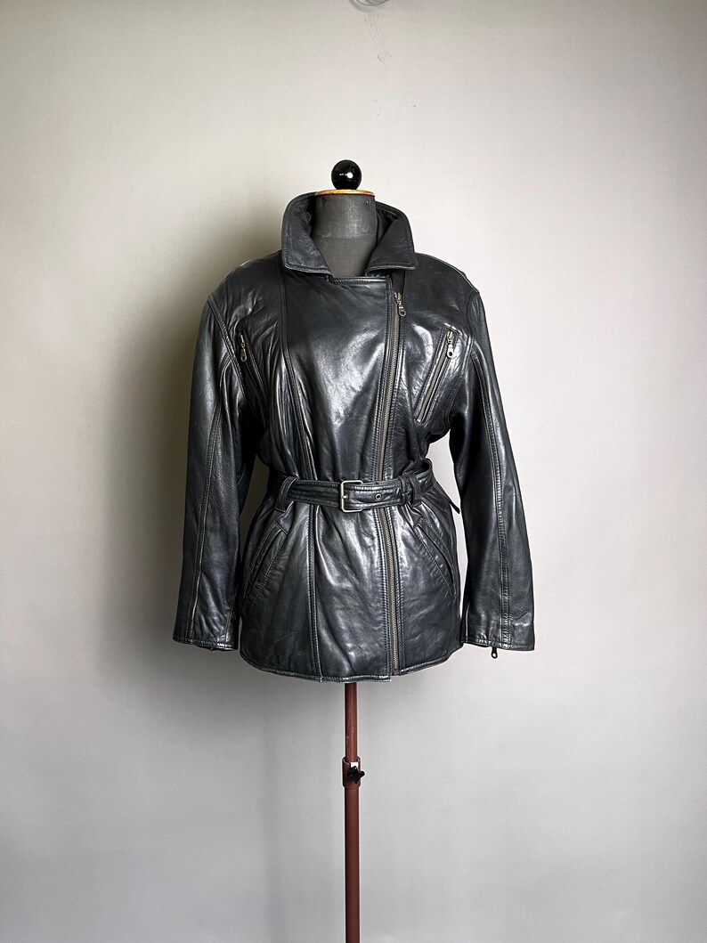 May include: A dark gray leather jacket with a belted waist and an asymmetrical zipper closure. The jacket features a wide collar, multiple zippered pockets, and a classic biker style. The jacket is displayed on a mannequin.