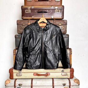 May include: A black leather jacket with a zip-up front, two front pockets, and a collared neckline. The jacket is hanging on a wooden hanger. The jacket is displayed on a stack of vintage suitcases.