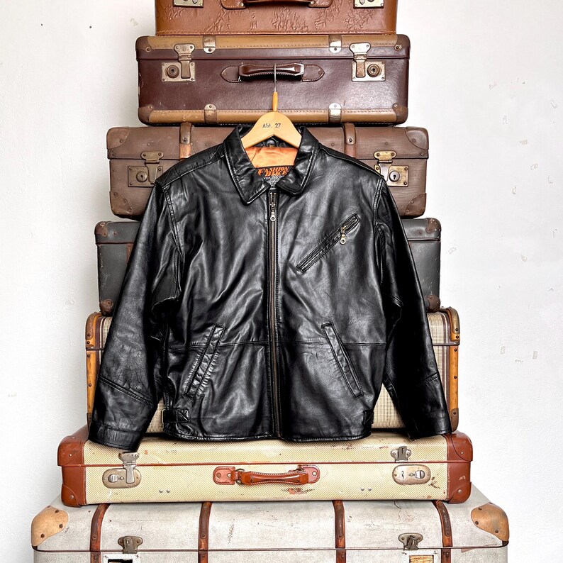 May include: A black leather jacket with a zip-up front, two front pockets, and a collared neckline. The jacket is hanging on a wooden hanger. The jacket is displayed on a stack of vintage suitcases.