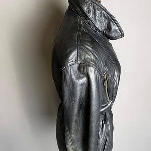 May include: Black leather jacket with a high collar and a belted waist. The jacket features a zipper closure and a sleek, vintage design. The leather appears worn, adding to its classic appeal.
