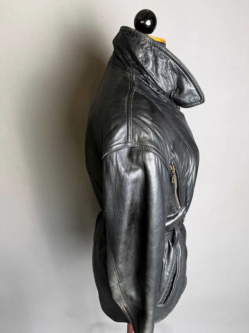 May include: Black leather jacket with a high collar and a belted waist. The jacket features a zipper closure and a sleek, vintage design. The leather appears worn, adding to its classic appeal.