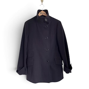 May include: A dark navy blue coat with a high collar, featuring a front button closure with four visible buttons, long sleeves with folded cuffs, and two rectangular flap pockets on the sides. The coat is displayed on a wooden hanger.