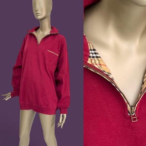 May include: A long-sleeve pullover sweatshirt in a deep red color featuring a quarter-zip neckline. The inner collar and the interior of the zipper placket are lined with a tan, black, and red plaid pattern. The sweatshirt has a small chest pocket.