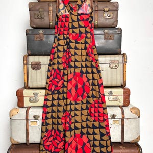 May include: A sleeveless maxi dress featuring a geometric pattern of black, tan, and bright red semicircles and circles. The dress has a V-neckline with wide shoulder straps that cross at the back, creating a fitted bodice that flows into a long skirt.