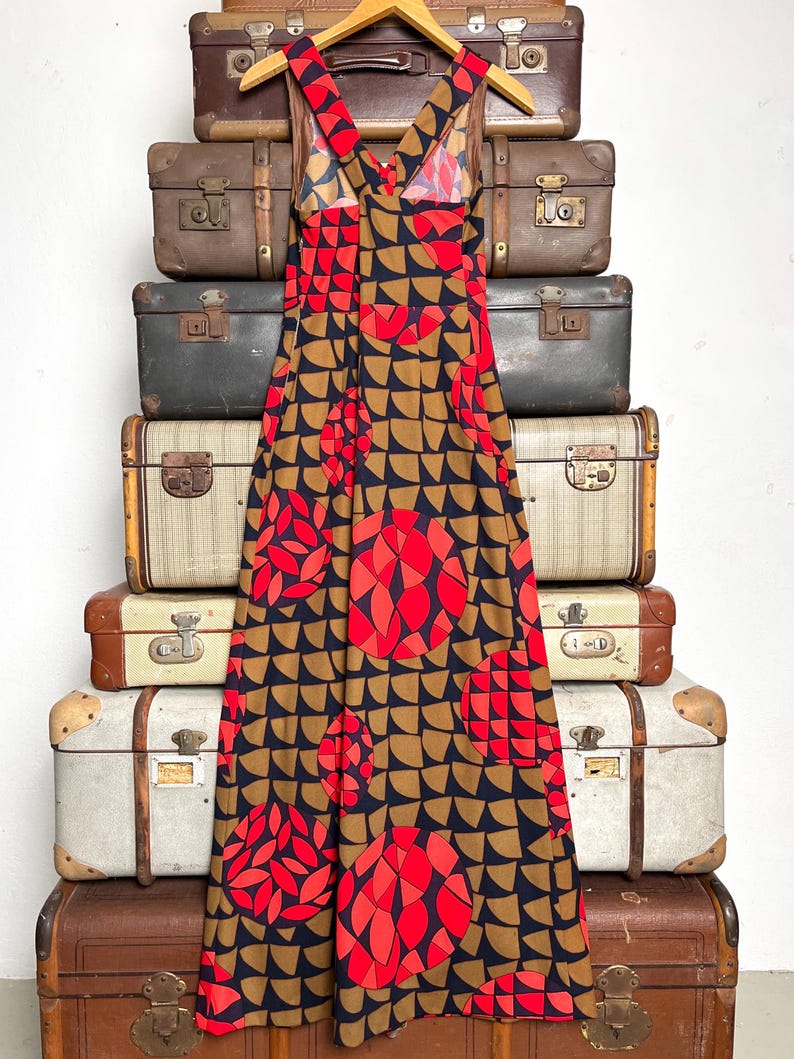 May include: A sleeveless maxi dress featuring a geometric pattern of black, tan, and bright red semicircles and circles. The dress has a V-neckline with wide shoulder straps that cross at the back, creating a fitted bodice that flows into a long skirt.