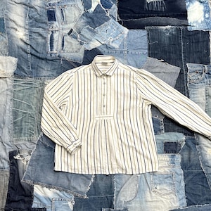 May include: A long-sleeved, button-down shirt with vertical stripes in shades of beige and blue. The shirt has a classic collar and a placket with three buttons. The shirt is displayed on a patchwork denim background.