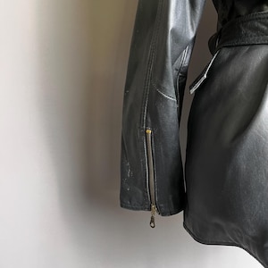 May include: Black leather jacket with a zipper detail on the sleeve. The jacket has a belt and a classic design. The leather appears worn, adding to its vintage appeal. The jacket is a medium weight.