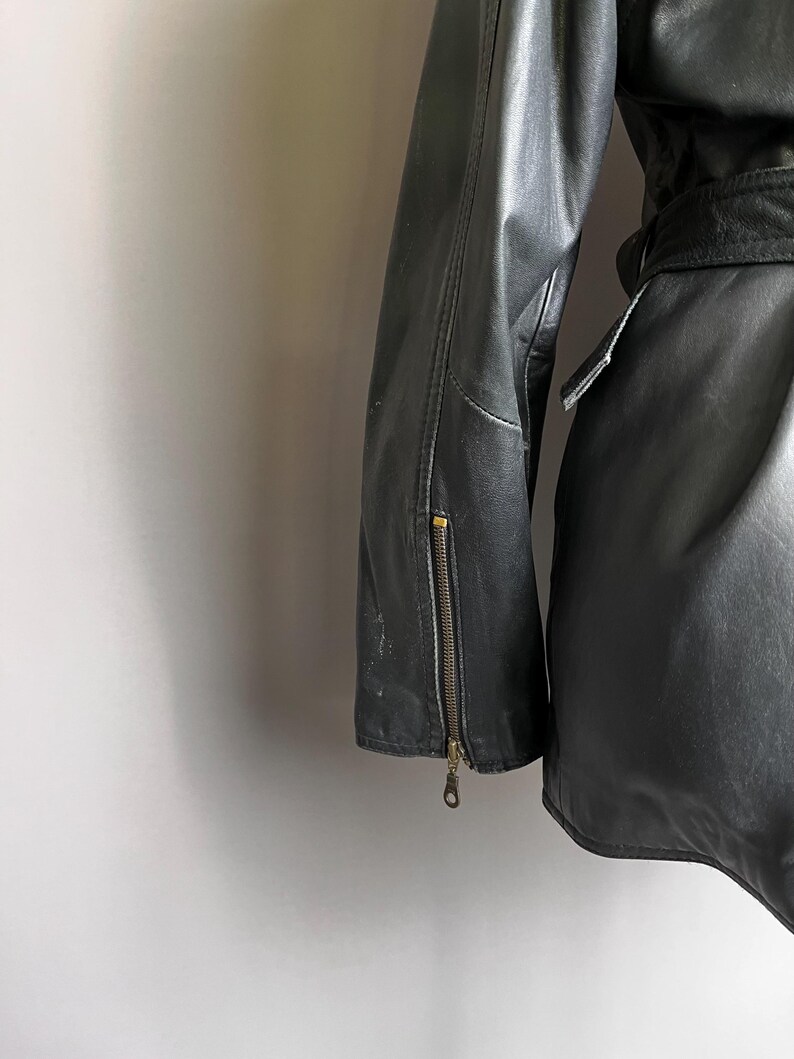 May include: Black leather jacket with a zipper detail on the sleeve. The jacket has a belt and a classic design. The leather appears worn, adding to its vintage appeal. The jacket is a medium weight.