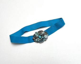 Könnte beinhalten: Ein türkisfarbener Gummibandgürtel mit einer großen, verzierten, blauen und goldenen Blumenschnalle.