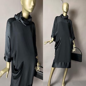 May include: A black dress with a cowl neck and three-quarter sleeves. The dress features satin accents on the sleeves, pockets, and hem. A black handbag is held by the mannequin. The dress is displayed on a mannequin with a gold-colored head and arms.