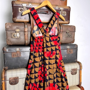May include: A sleeveless dress featuring a repeating geometric pattern of semicircles and circles in shades of red, brown, and black. The dress has a V-neckline with wide shoulder straps that cross at the back and a fitted bodice that flares.