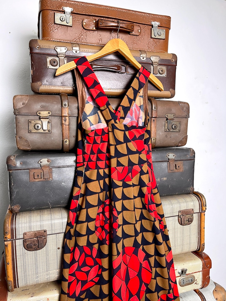May include: A sleeveless dress featuring a repeating geometric pattern of semicircles and circles in shades of red, brown, and black. The dress has a V-neckline with wide shoulder straps that cross at the back and a fitted bodice that flares.