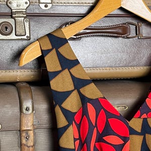 May include: A sleeveless dress featuring a dark navy blue base with a geometric pattern of tan semi-circles on the shoulder straps and a vibrant red leaf-like print on the bodice, displayed on a light-colored wooden clothes hanger.