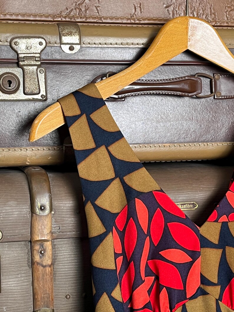 May include: A sleeveless dress featuring a dark navy blue base with a geometric pattern of tan semi-circles on the shoulder straps and a vibrant red leaf-like print on the bodice, displayed on a light-colored wooden clothes hanger.