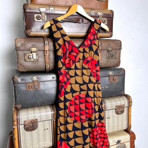 May include: A sleeveless dress featuring a V-neckline and a geometric pattern of black, tan, and red semicircles. The fabric displays a repeating motif of large circular shapes composed of smaller red and black semicircles against a tan background.