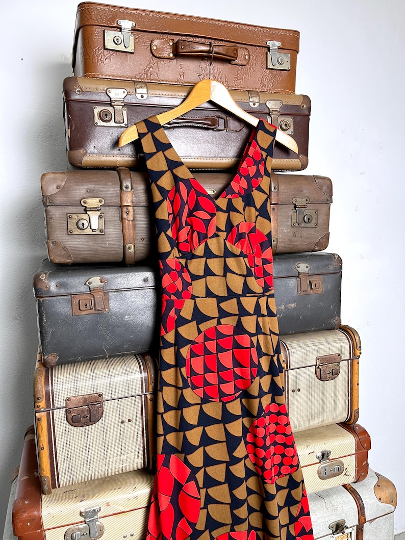 May include: A sleeveless dress featuring a V-neckline and a geometric pattern of black, tan, and red semicircles. The fabric displays a repeating motif of large circular shapes composed of smaller red and black semicircles against a tan background.
