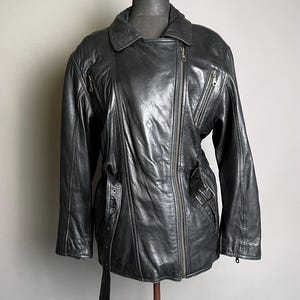 May include: Black leather jacket with an asymmetrical zipper closure, multiple zippered pockets, and a belt detail at the waist. The jacket has a classic biker style with a notched collar and long sleeves. The jacket is displayed on a mannequin.