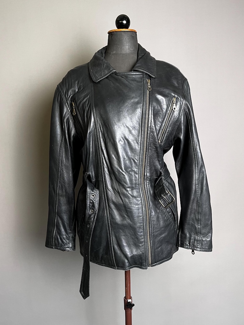 May include: Black leather jacket with an asymmetrical zipper closure, multiple zippered pockets, and a belt detail at the waist. The jacket has a classic biker style with a notched collar and long sleeves. The jacket is displayed on a mannequin.