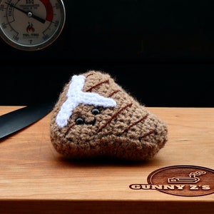 May include: A brown crocheted steak with a white fat cap and a cute face sits on a wooden cutting board with a black handled knife. The cutting board has the text "Gunny's BBQ" burned into the wood.