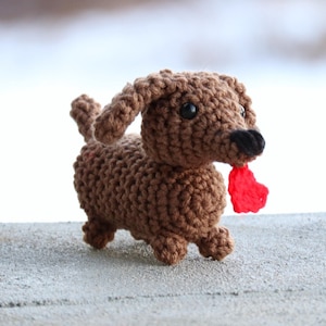 May include: A brown crocheted dachshund toy holding a red heart in its mouth. The toy is standing on a gray concrete surface.
