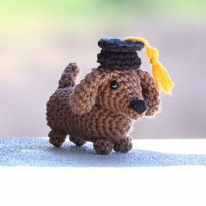 May include: A small brown crocheted dachshund figurine wearing a black graduation cap with a yellow tassel. The dog has dark eyes, a black nose, and floppy ears, crafted with a textured yarn stitch pattern in a standing position.