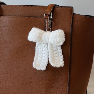 May include: A white, crocheted bow-shaped keychain hanging from a brown leather handbag. The bow is made of a textured, off-white yarn and is attached to a silver-colored clasp.