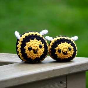 May include: Two handmade crochet bee toys. The bees are yellow and black with white wings and smiling faces. They are sitting on a wooden surface with a green background.