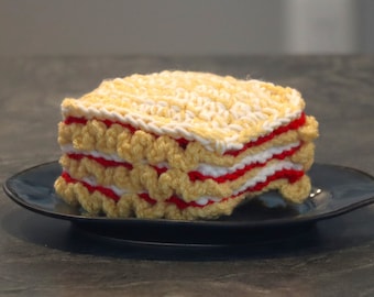 May include: A crocheted cake made of yellow, white, and red yarn. The cake is sitting on a dark blue plate.