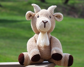 May include: A crocheted goat plush toy with brown hooves, a white beard, and brown eyes. The goat is sitting on a wooden deck with a green grassy background.