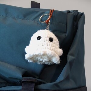 May include: A white crocheted ghost keychain with black button eyes and a ruffled bottom. The keychain is attached to a silver ring and a brown leather strap. The ghost is hanging on a teal blue bag.