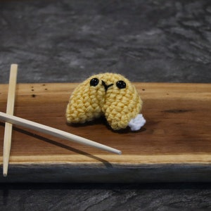 May include: A handmade, crocheted fortune cookie with black eyes and a white accent, resting on a wooden surface. Two wooden chopsticks are crossed beside the cookie. The cookie is yellow and appears to be made of yarn.