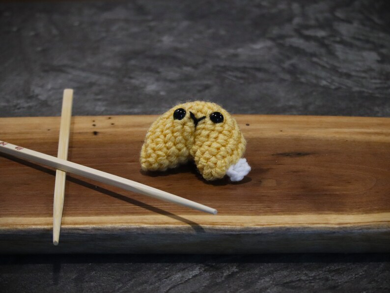 May include: A handmade, crocheted fortune cookie with black eyes and a white accent, resting on a wooden surface. Two wooden chopsticks are crossed beside the cookie. The cookie is yellow and appears to be made of yarn.
