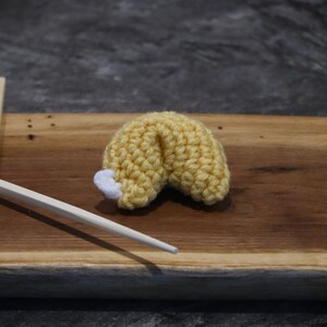 May include: A crocheted fortune cookie in yellow with a white tip, resting on a wooden board. Two wooden chopsticks are placed to the left of the cookie. The background is a dark gray.