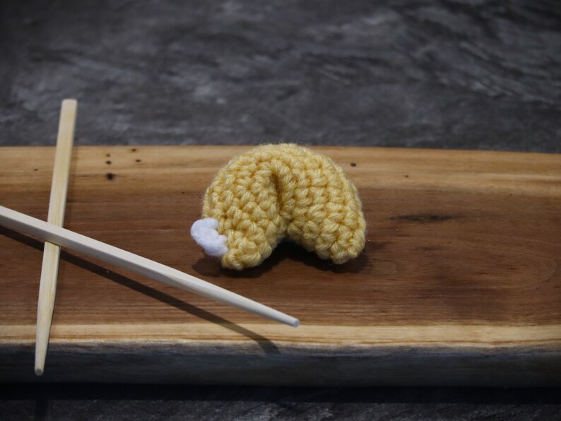 May include: A crocheted fortune cookie in yellow with a white tip, resting on a wooden board. Two wooden chopsticks are placed to the left of the cookie. The background is a dark gray.