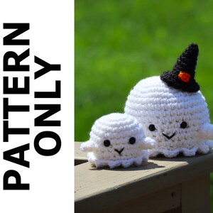 May include: Two crocheted ghost ornaments, one wearing a black witch hat with an orange stripe. The larger ghost has a happy face with black eyes and a smile. The smaller ghost has a similar face. Both ghosts are white and are sitting on a wooden surface.