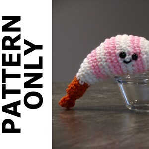 May include: A crocheted shrimp amigurumi toy with pink and white stripes, orange legs, and black eyes. The toy is smiling and is sitting on a clear glass bowl.