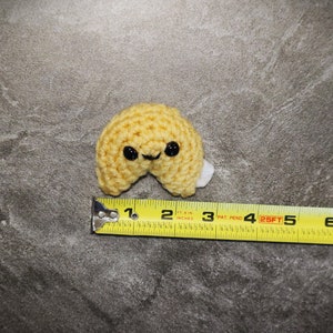 May include: A crocheted fortune cookie in a light yellow color with black button eyes and a small white paper piece. The cookie measures approximately 2.5 inches in length. The background is a mottled gray surface.