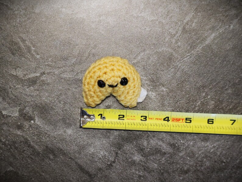 May include: A crocheted fortune cookie in a light yellow color with black button eyes and a small white paper piece. The cookie measures approximately 2.5 inches in length. The background is a mottled gray surface.