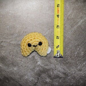 May include: A crocheted fortune cookie in yellow yarn with black eyes and a smiling mouth. The cookie is approximately 2 inches (5 cm) wide. A white piece of fabric is visible inside the cookie. The item is on a gray surface.
