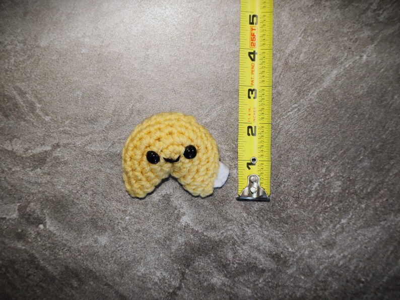 May include: A crocheted fortune cookie in yellow yarn with black eyes and a smiling mouth. The cookie is approximately 2 inches (5 cm) wide. A white piece of fabric is visible inside the cookie. The item is on a gray surface.