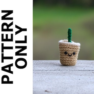 May include: A crocheted coffee cup with a green straw and a smiling face. The cup is beige and white and has a brown lid. The cup is sitting on a light grey surface.